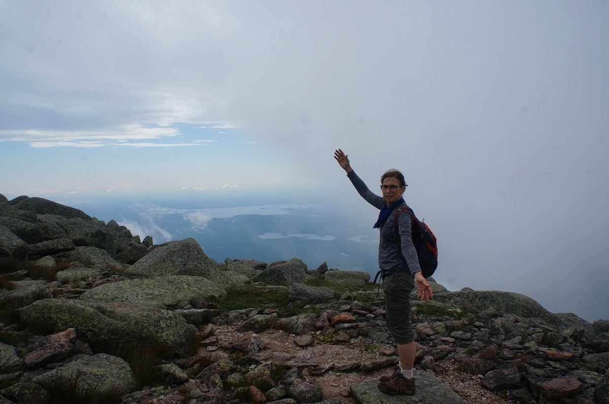 A woman raises her arm on a mountain with clouds in the background.