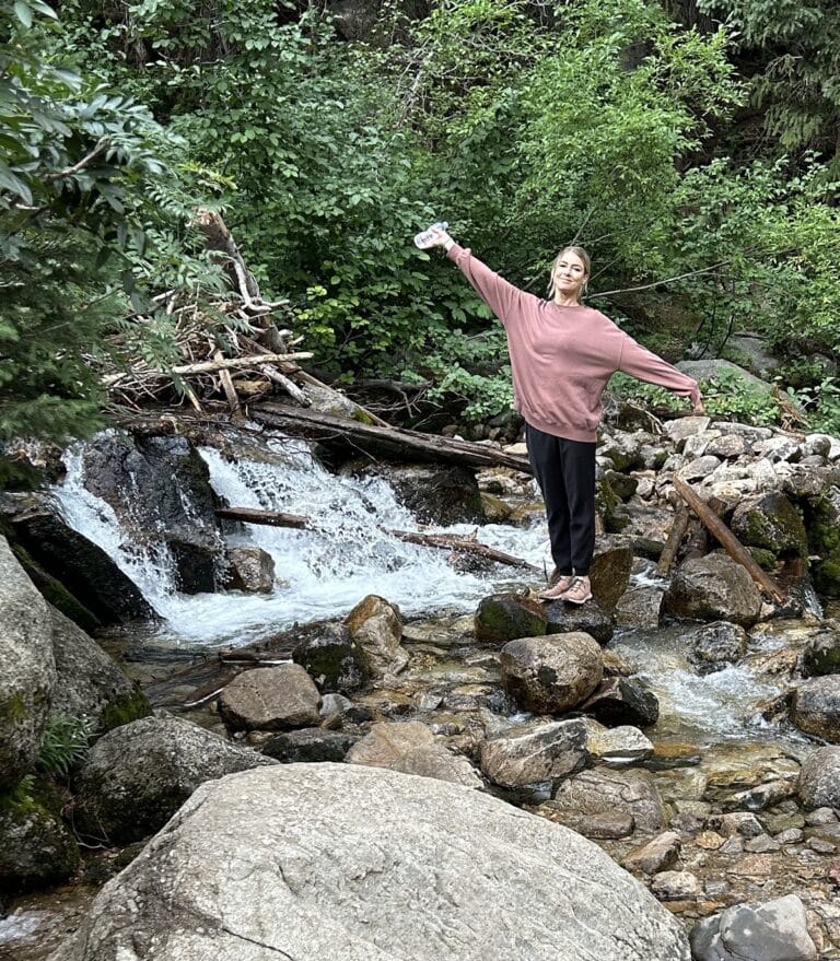A woman stretches her arm out with a waterfall behind her.