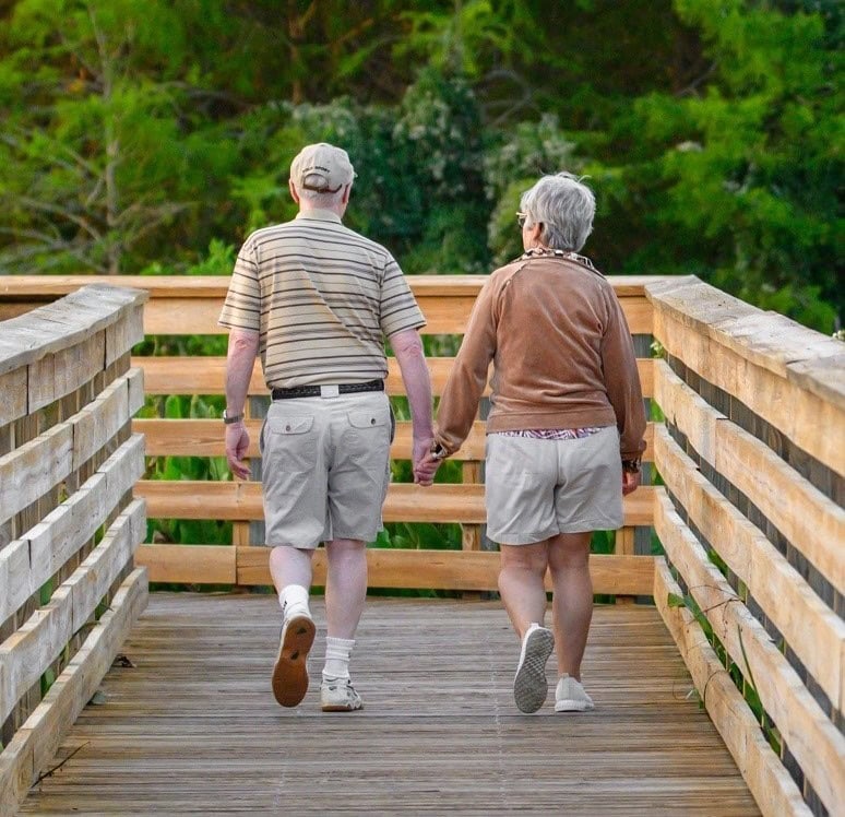 An older manand woman walk along a boardwalk holding hands.