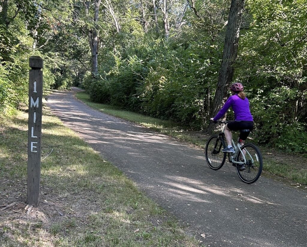 A woman rides a bike on a trail passing a 1-mile marker.