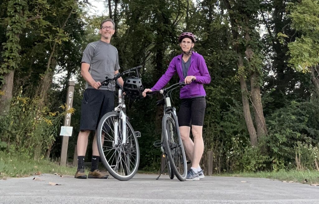 A man and woman smile for a picture holding onto their bikes at a trailhead.