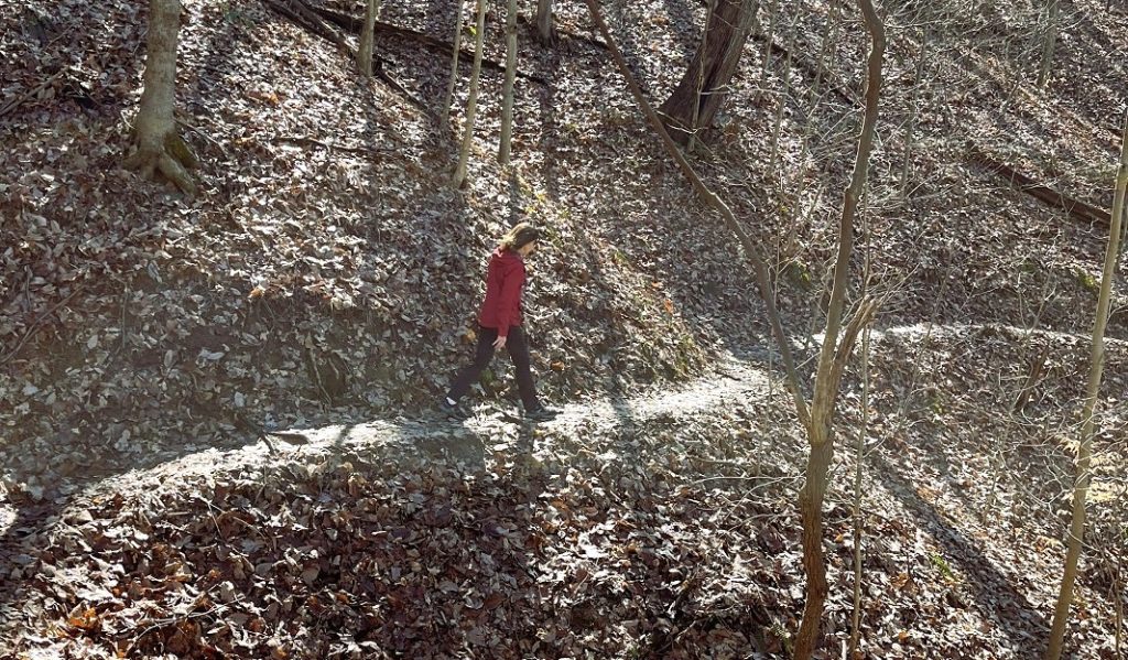 A woman in a bright jacket walks on a leafy path on the side of a hill.