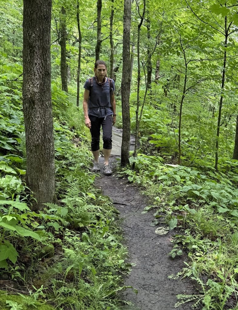 A woman walks a think dirt trail wearing a backpack.
