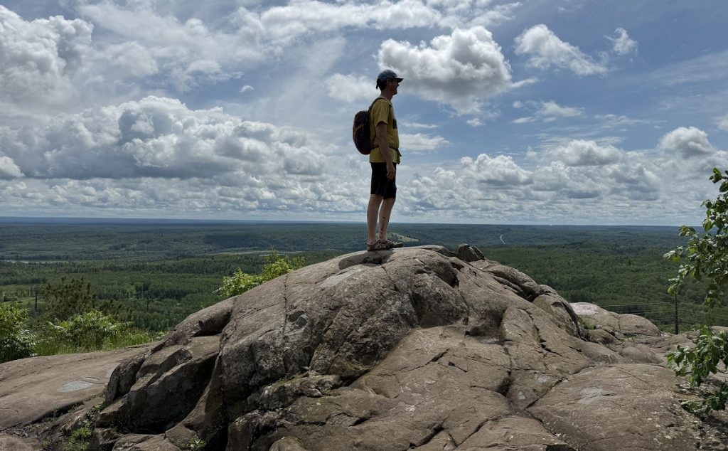A man stands on the top of a rocky peak with a cloudy sky behind him.