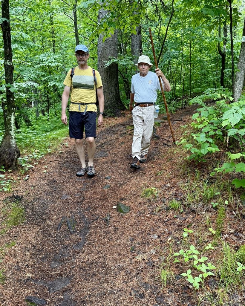 Two older men, one with a walking stick, walk side by side on a trail.