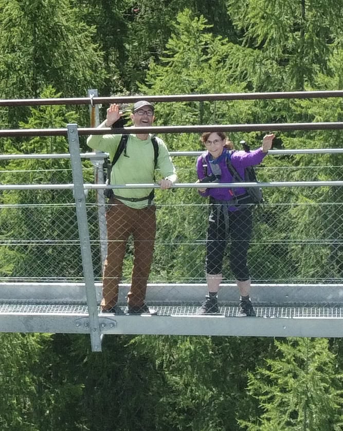 A man and a woman wave to a camera on a drone near them on the suspension bridge.