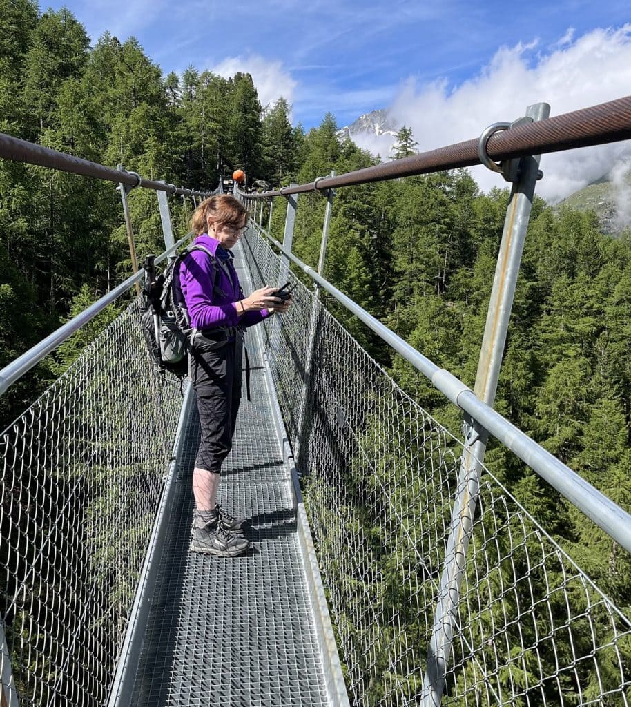 A woman holds a controller to fly her drone while standing on a suspension bridge.