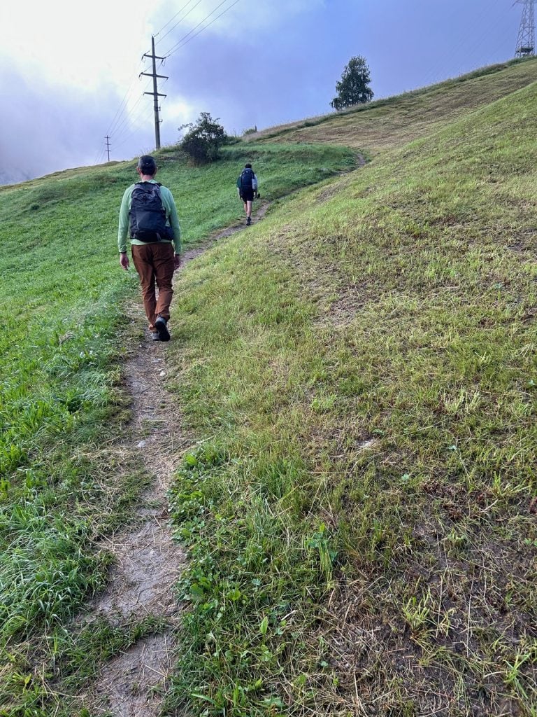 A man follows a hiker along a dirt path outside a town.