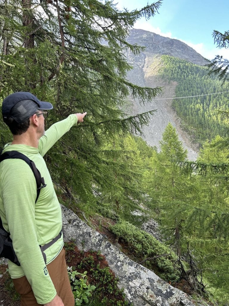A hiker with a hat points in the distance to a long suspension bridge.