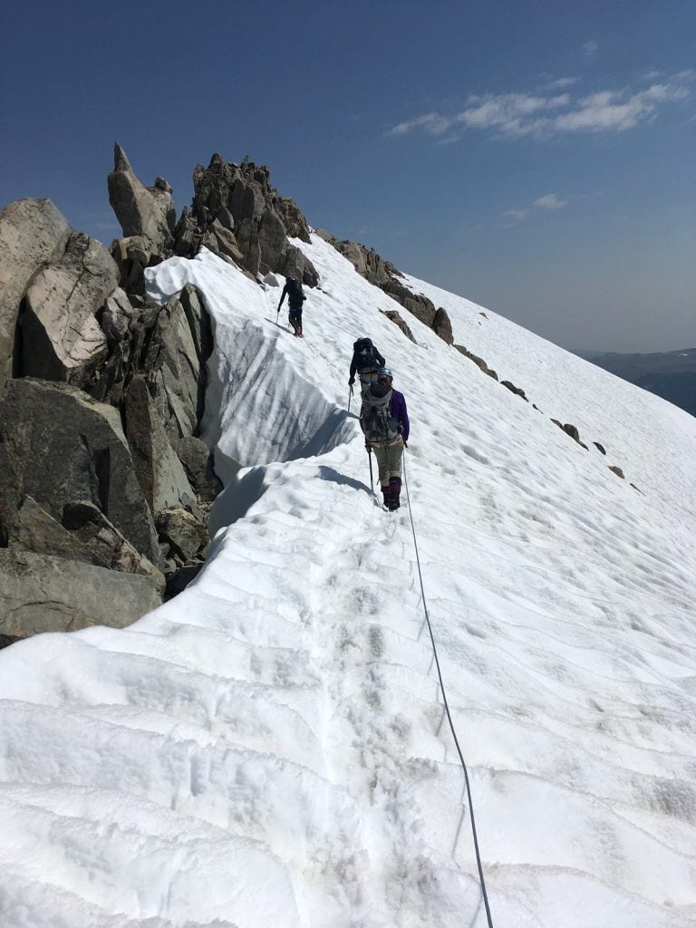 Climbers are roped up along a snowy ridge at the top of a  mountain.