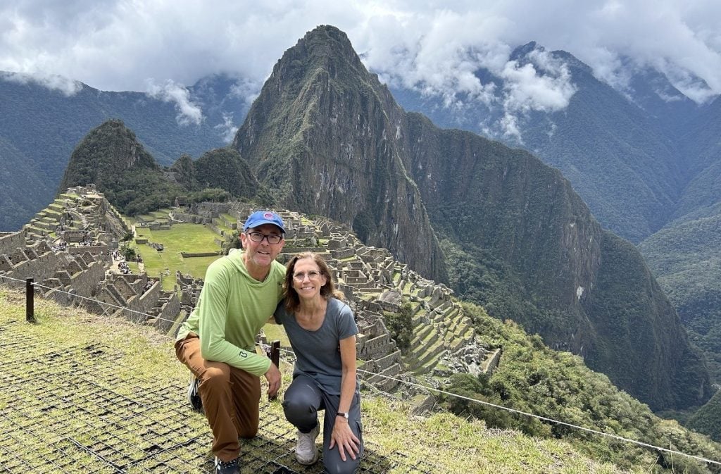 A man and woman crouch down in front of Machu Picchu and the big mountain behind it.