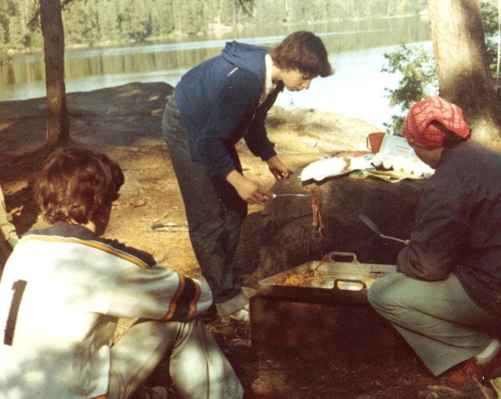 A teenage girl leans over a pan cooking outdoors at a campsite.