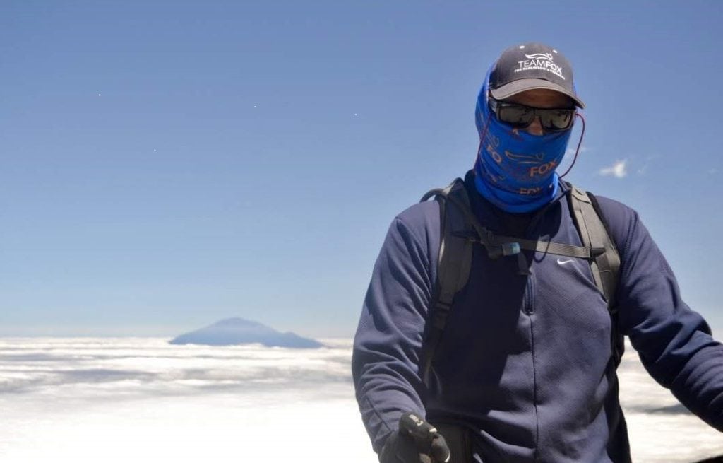 A man with his face covered stands on a snowy mountaintop.