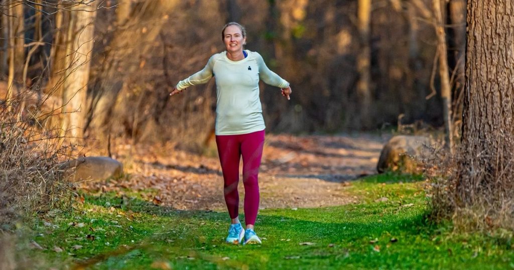 A woman walks through a wooded area with her arms outstretched.