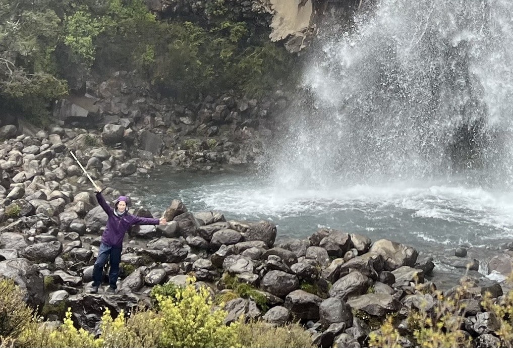 A woman raises her walking stick next to a waterfall.