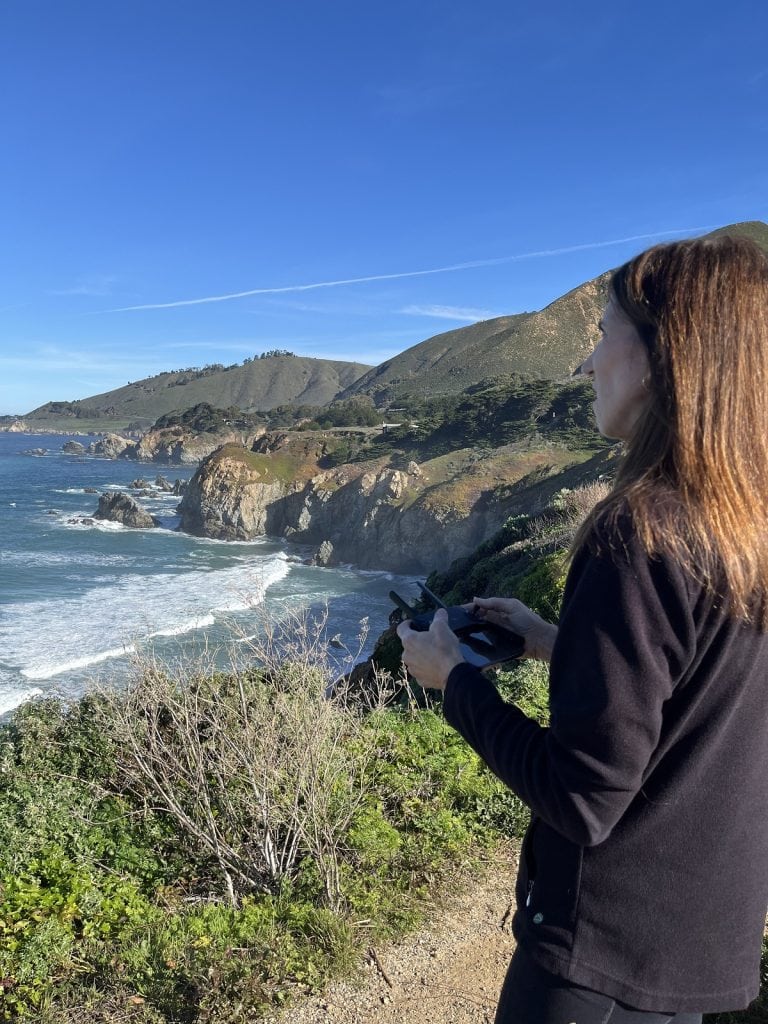 A woman holds a drone controller while flying it on the coast.