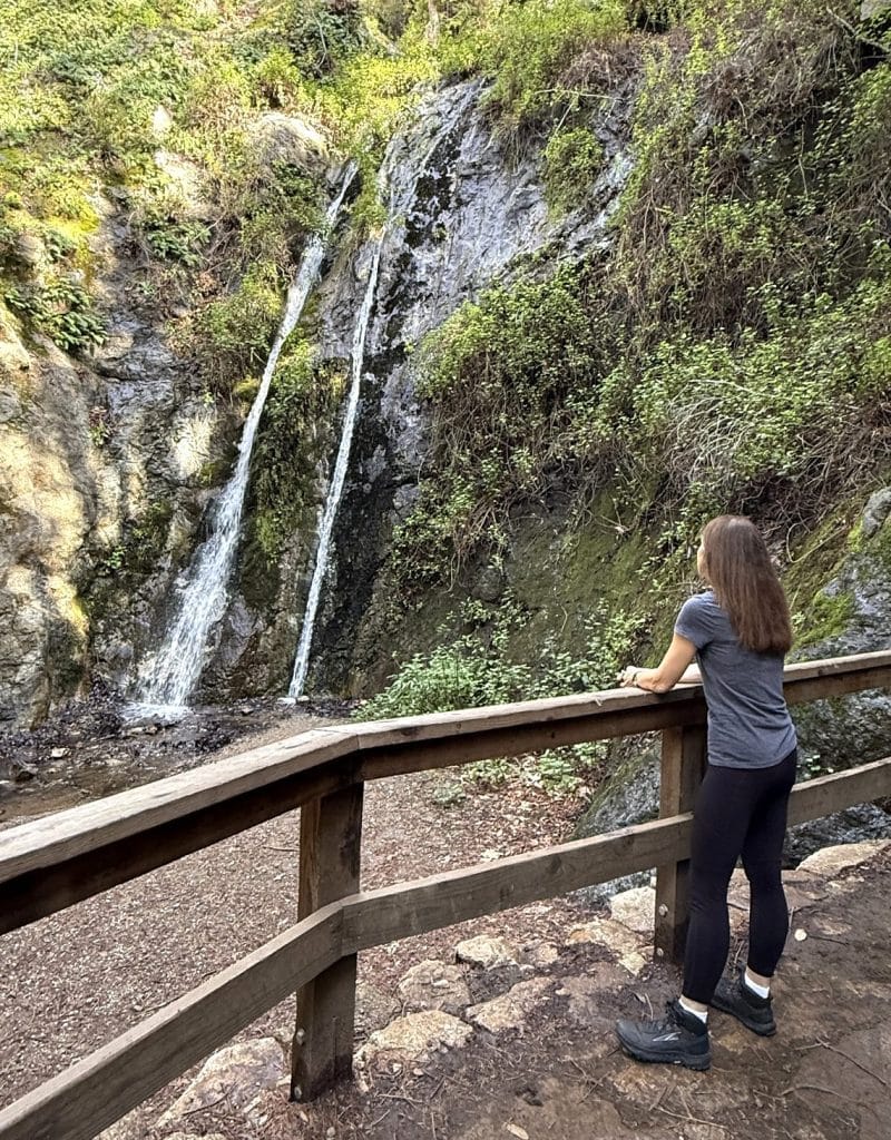 A woman stands at a wooden fence looking at a waterfall.