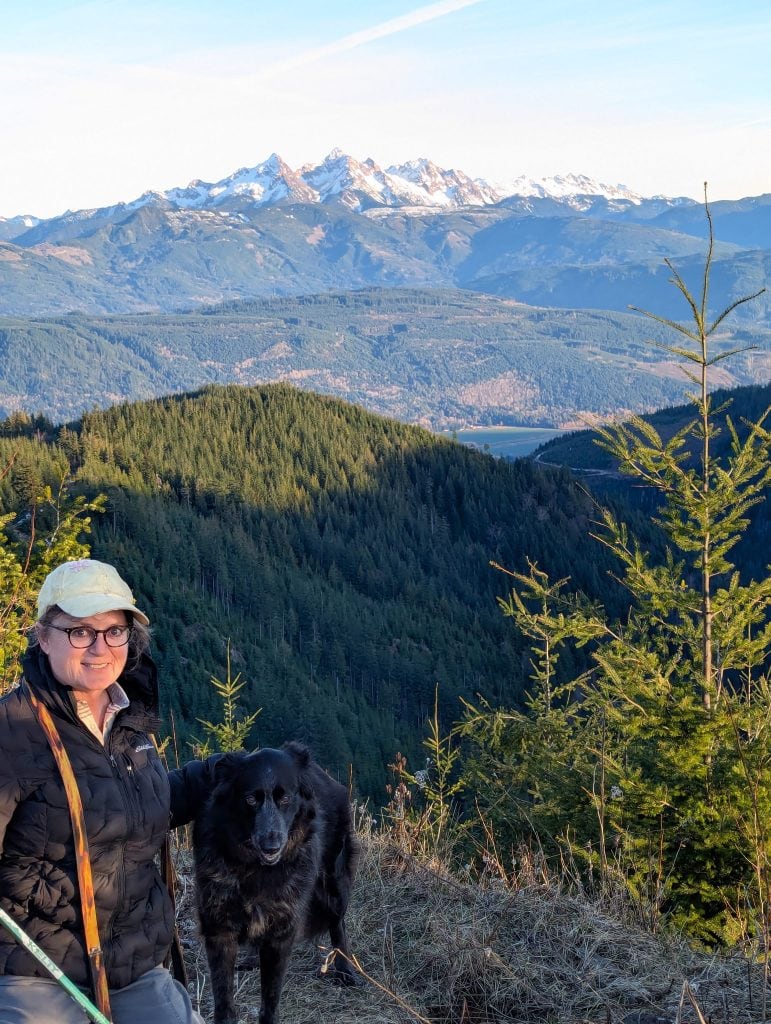 A woman and black dog stand before a scenic mountainscape.