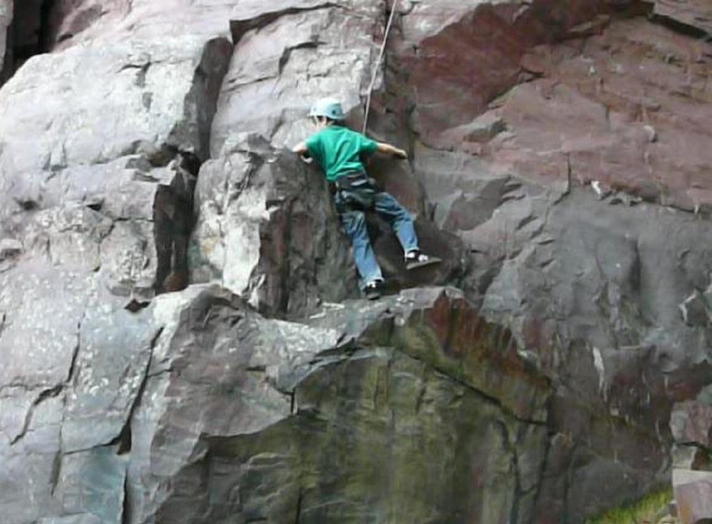 A boy clings to a rock while roped up for climbing.