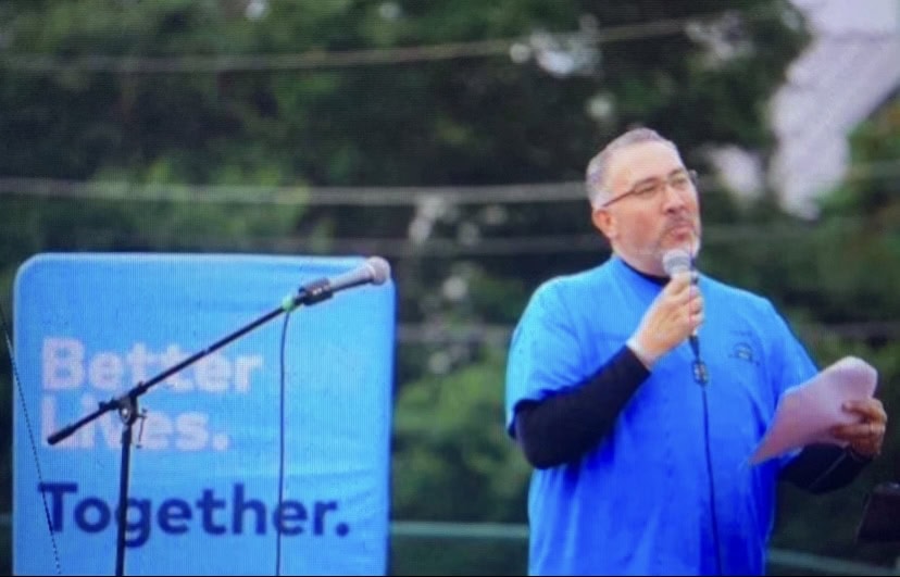 A man speaks in a microphone with a blue banner that says "Better Lives Together."