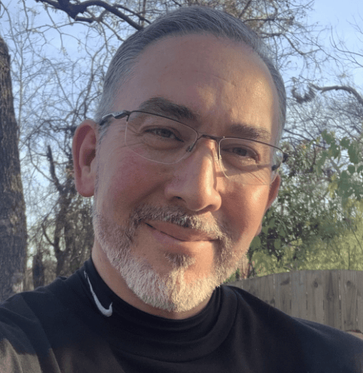 A man with glasses is smiling while outdoors.