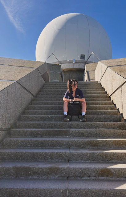 A woman sits on the stairs leading up to what looks like an observatory dome.
