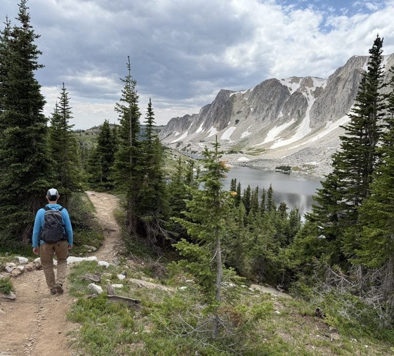 A man hikes past trees, a lake, and mountains.
