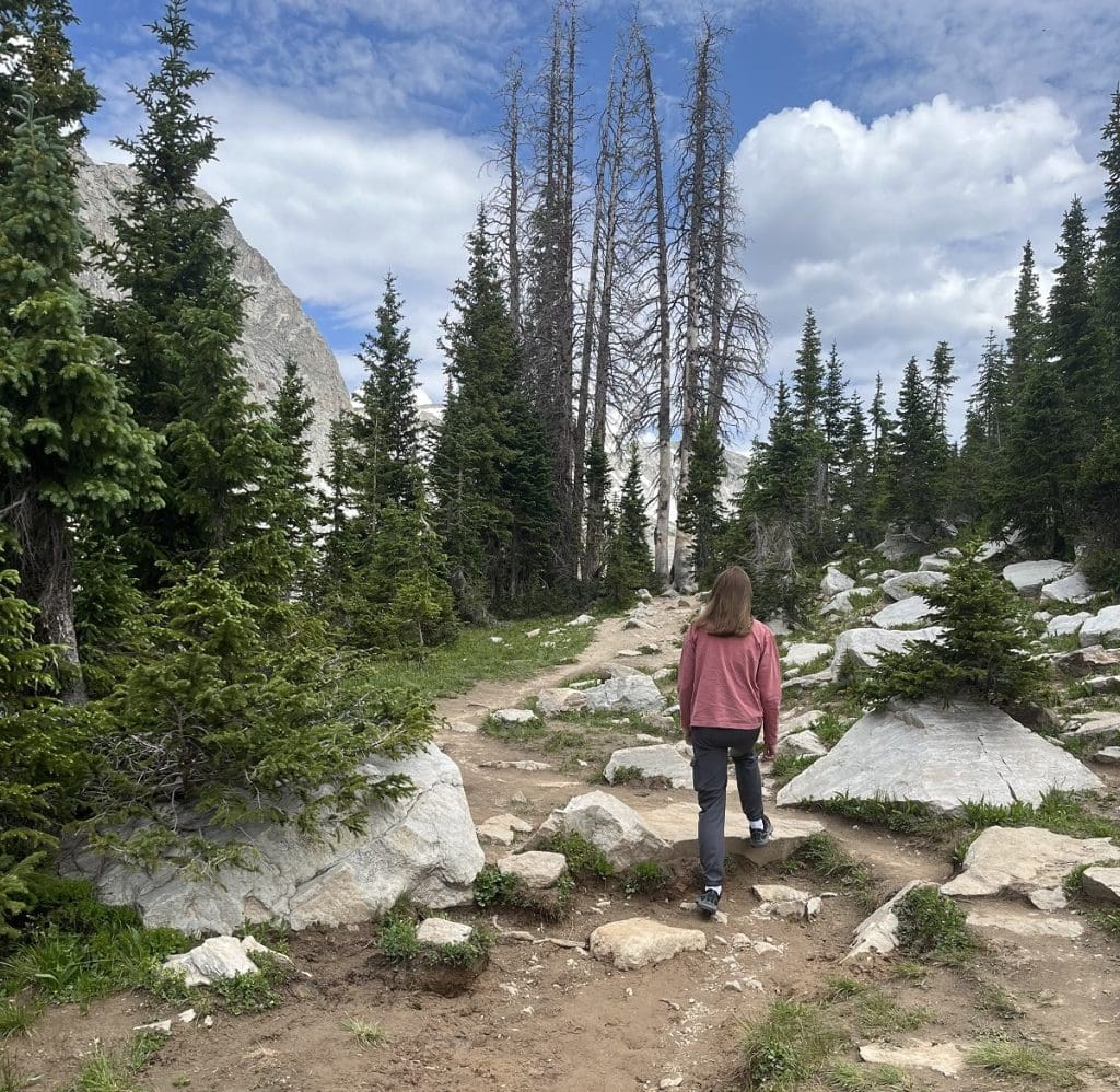 A woman in a red shirt is shown hiking a rocky path from behind.