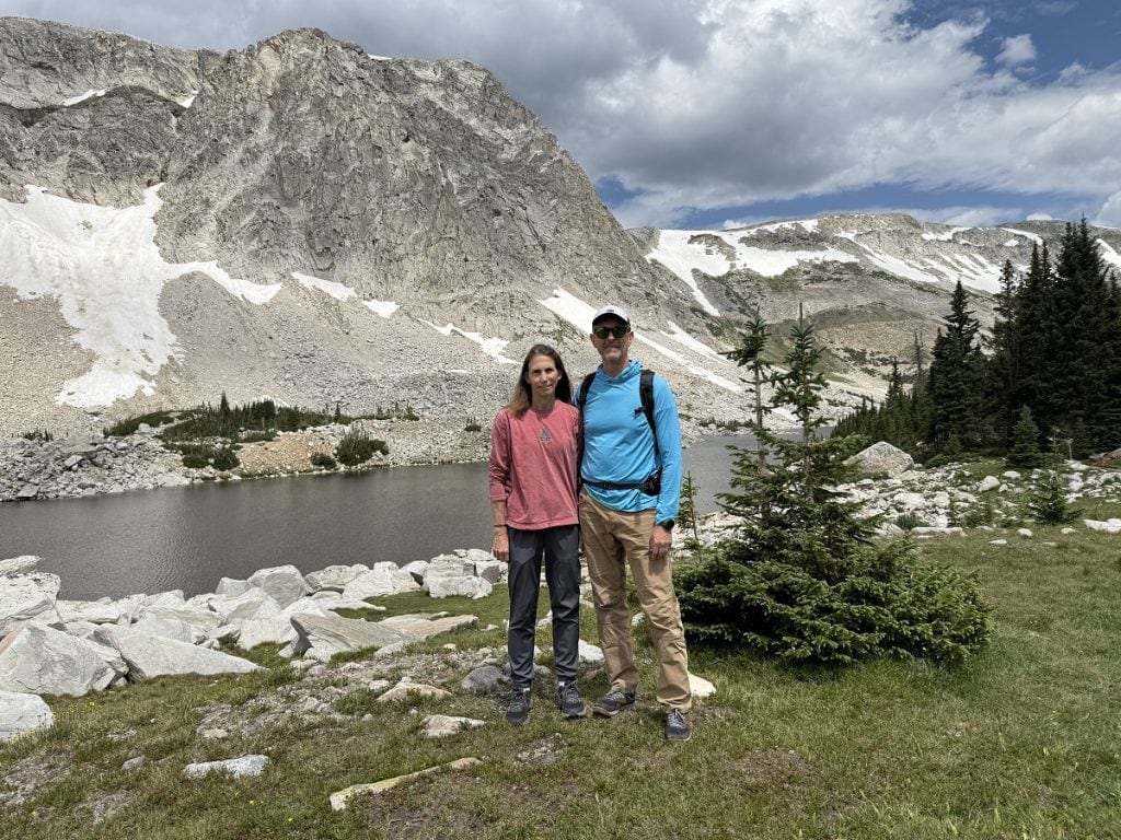 A man and woman, hikers, stand in front of a gray lake.