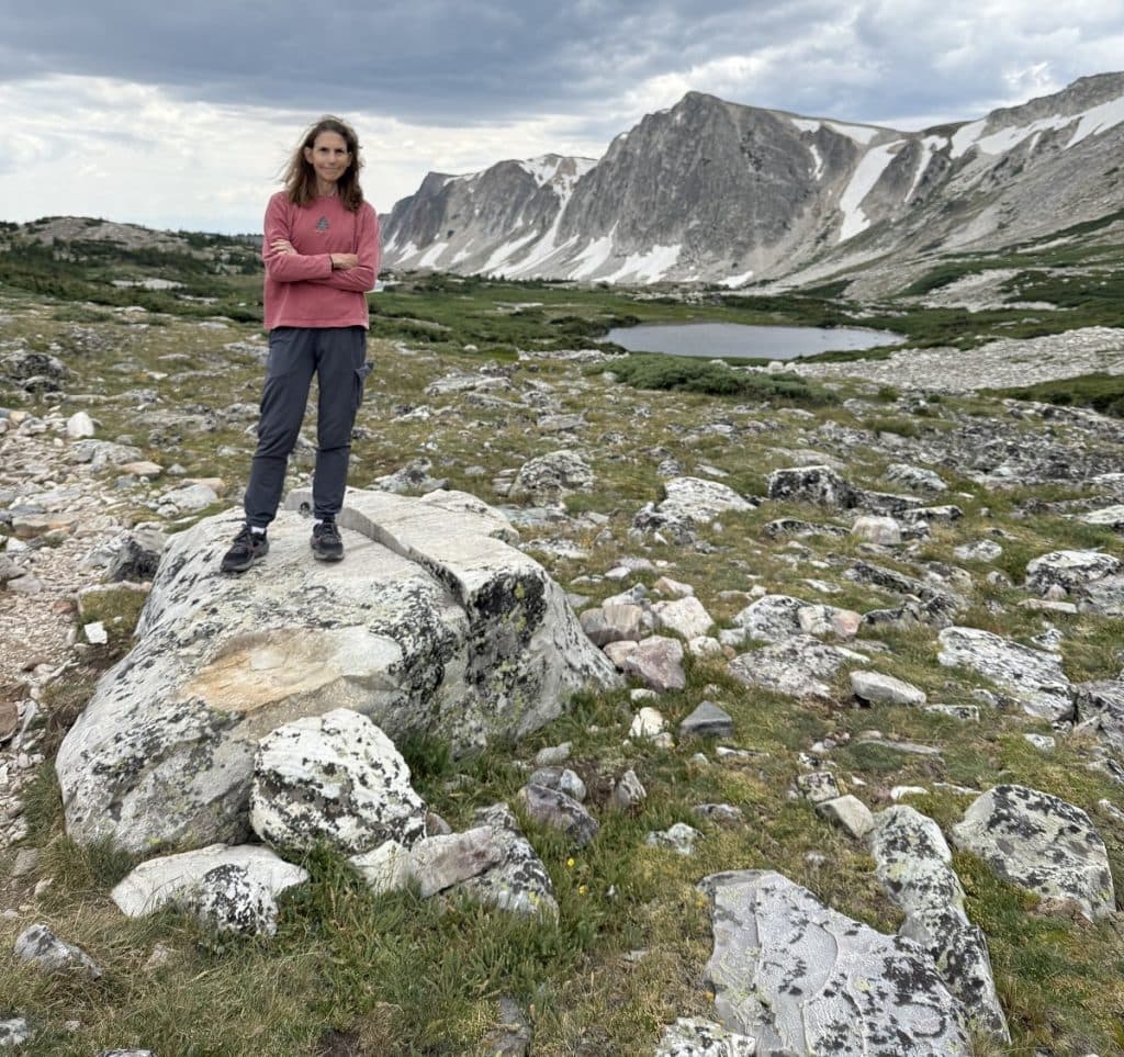 A woman in a red shirt stands on a big rock with her arms folded.