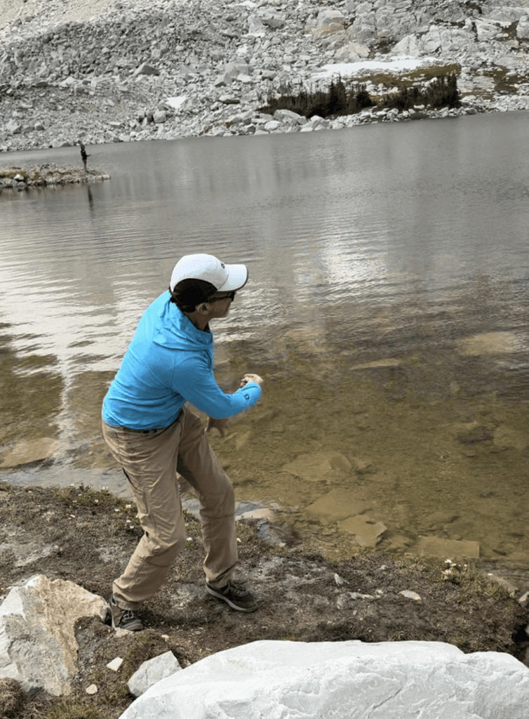 A man with a cap and blue shirt skips rocks into a lake.