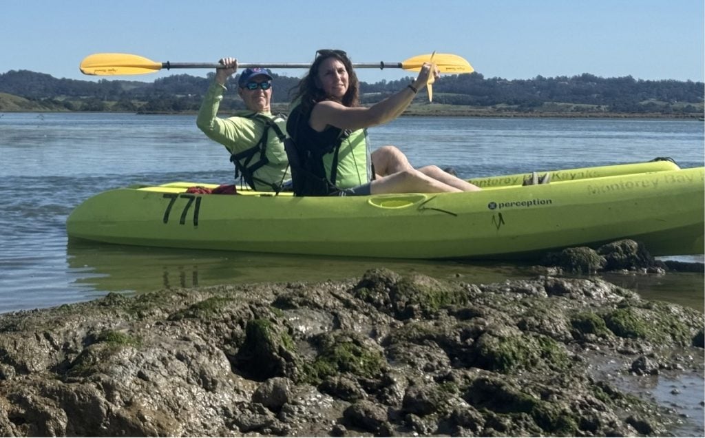 A man and woman hold up their paddles while parked in kayaks off the side of the waterway.