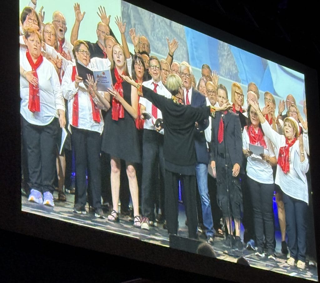 A group of men and women in red scarves sing on a stage.