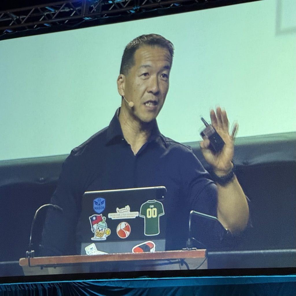 A man on a huge monitor speaks with his hand up.