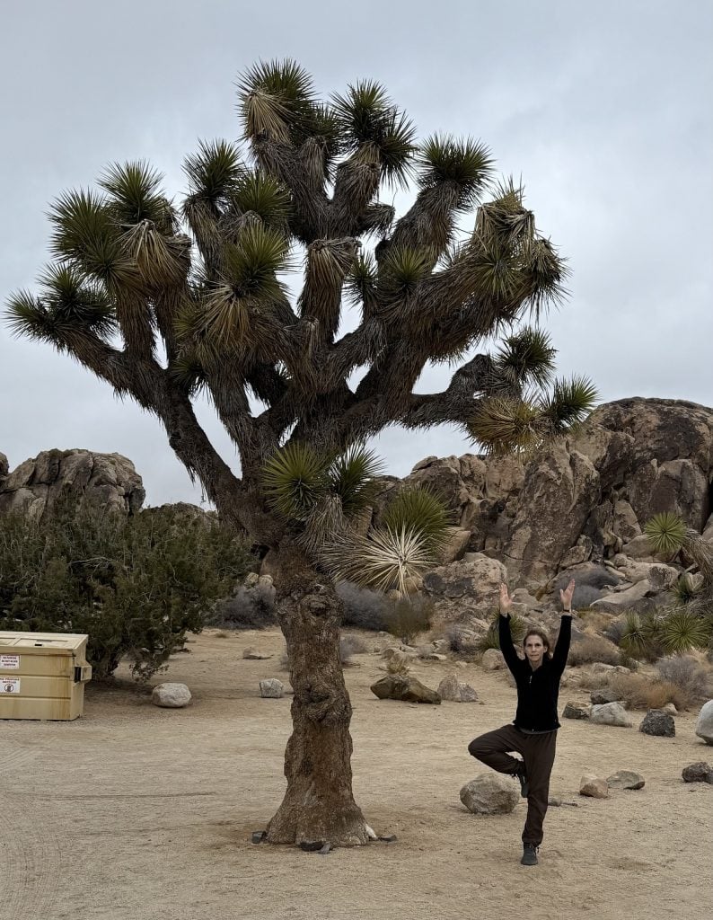 A woman stands in a tree pose with a natural background outside with a tree.