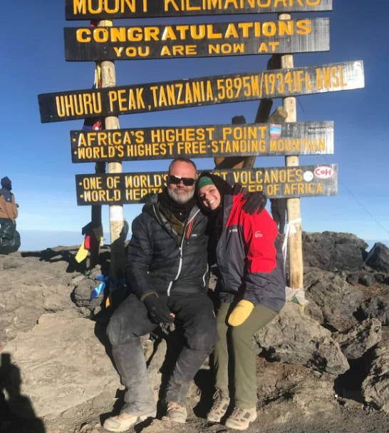 A man and his grown daughter sit under a sign at the top of Kilimanjaro.