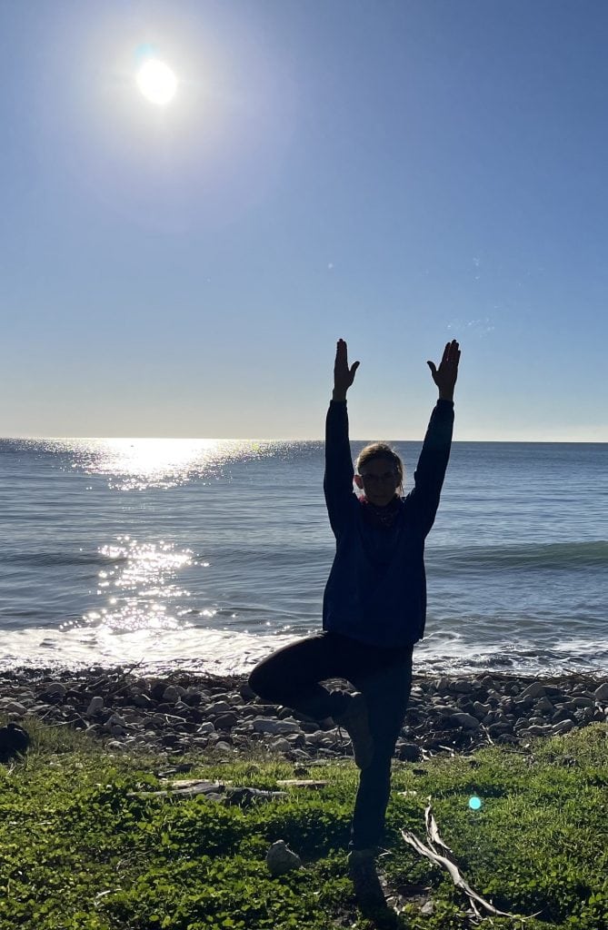 A woman stands in a tree pose with an ocean in the background outside.