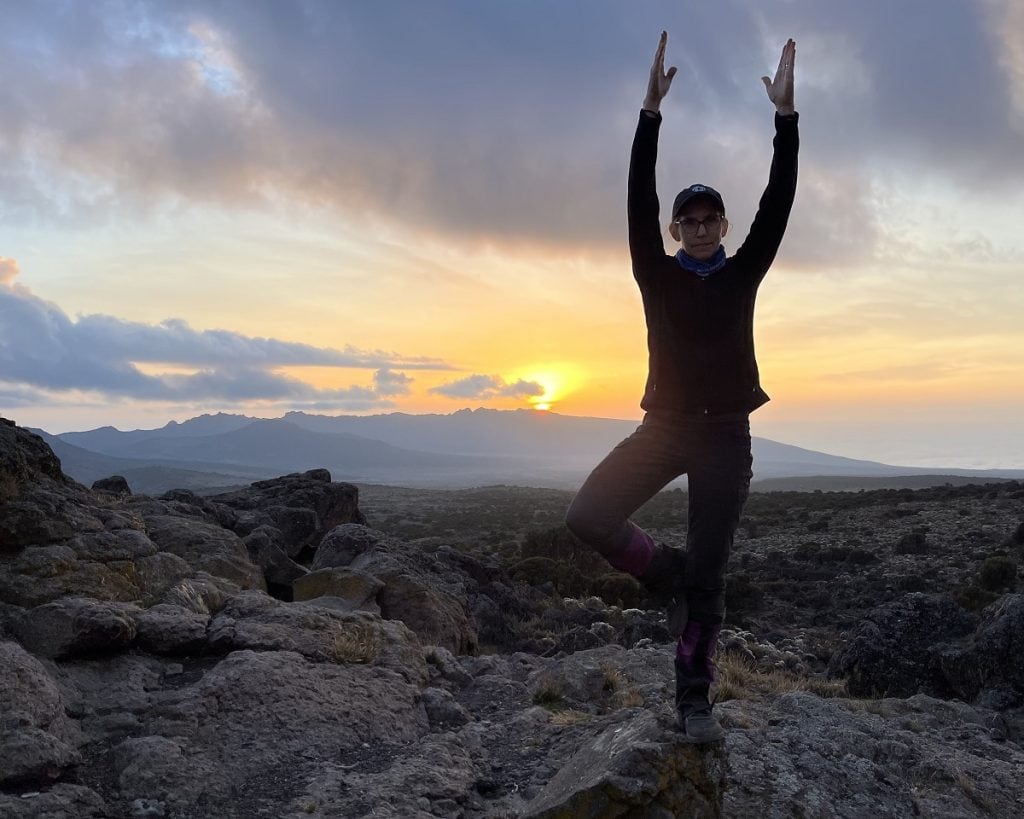 A woman stands in a tree pose with a natural background outside.