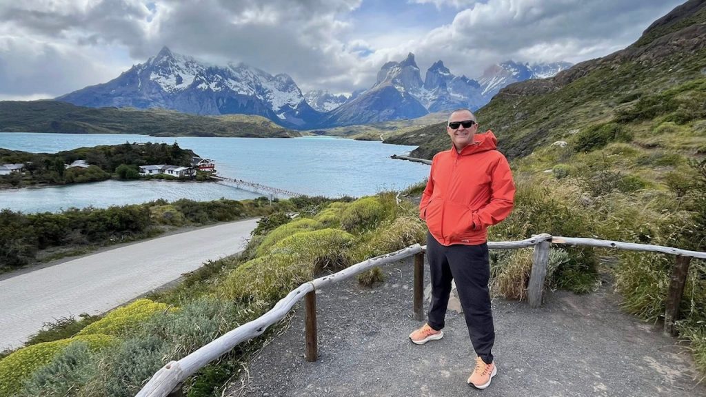 A hiker in an orange shirt stands in front of a mountain lake.
