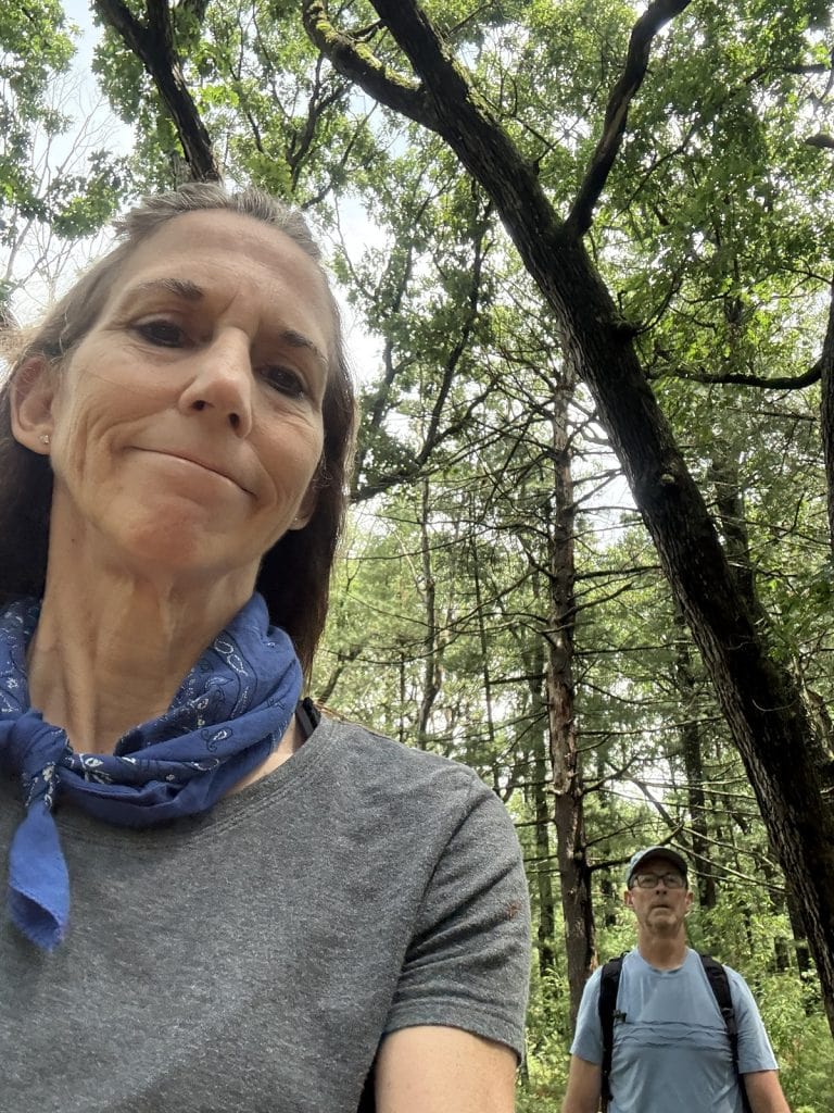 A woman hiker takes a selfie of herself and her husband in the background.