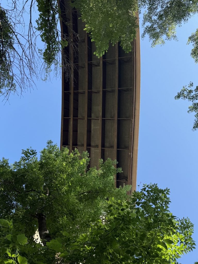 a large reddish metal piece is framed by trees and backed by blue sky.