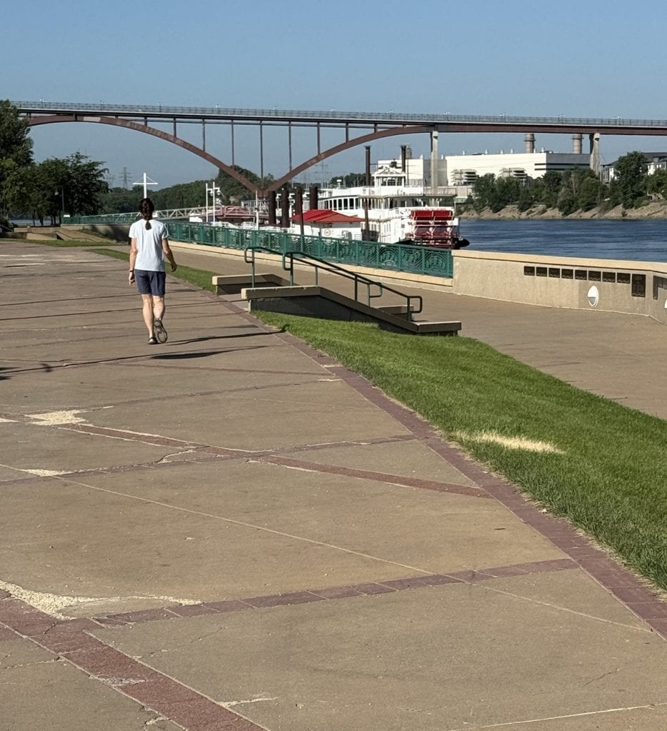 A woman walks along a concrete path with the river up ahead.